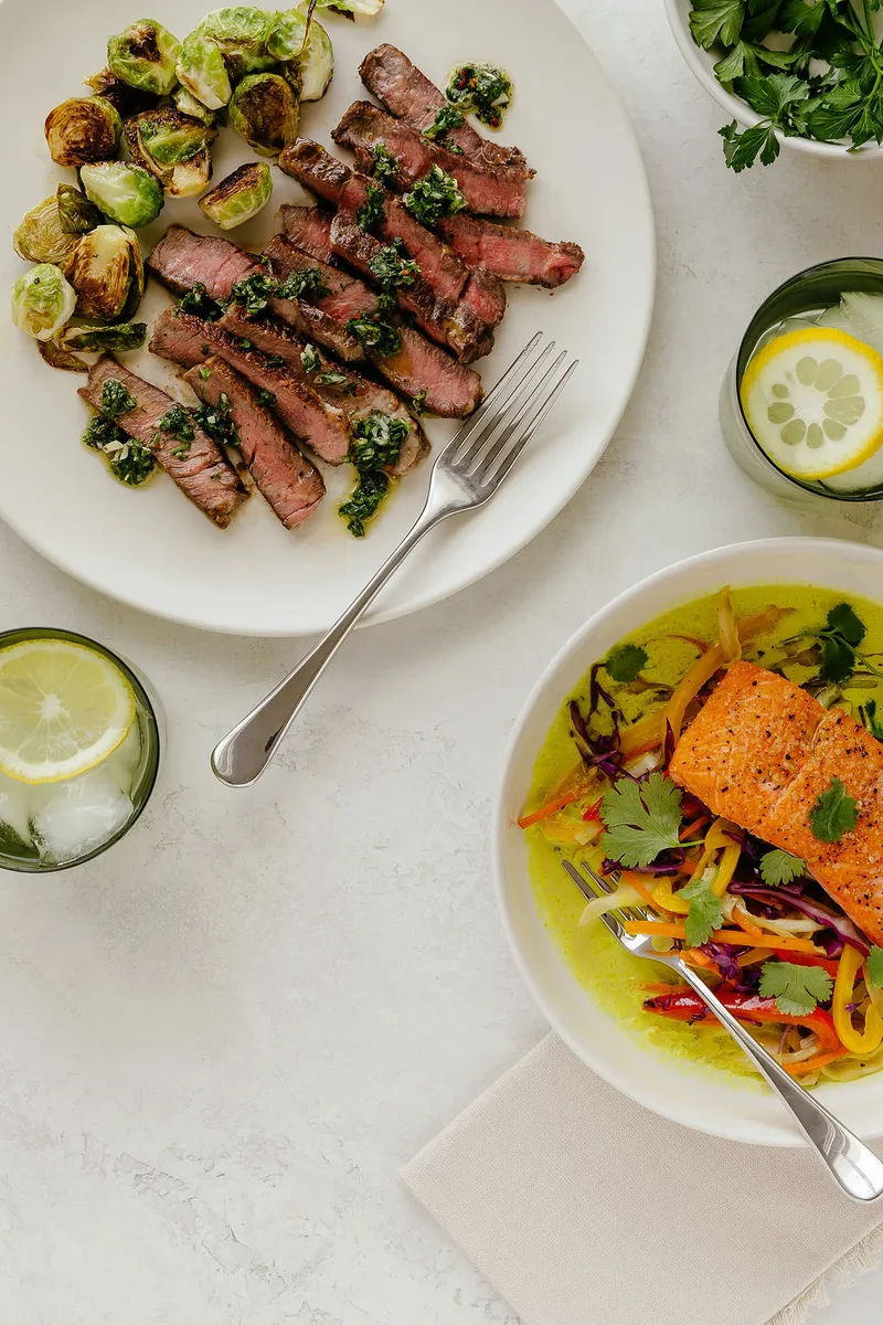 Two Glyvance meals — steak with chimichurri and salmon in curry sauce — plated with drinks and fresh herbs